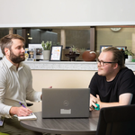 Two men speak seated at a table with a laptop open before them.