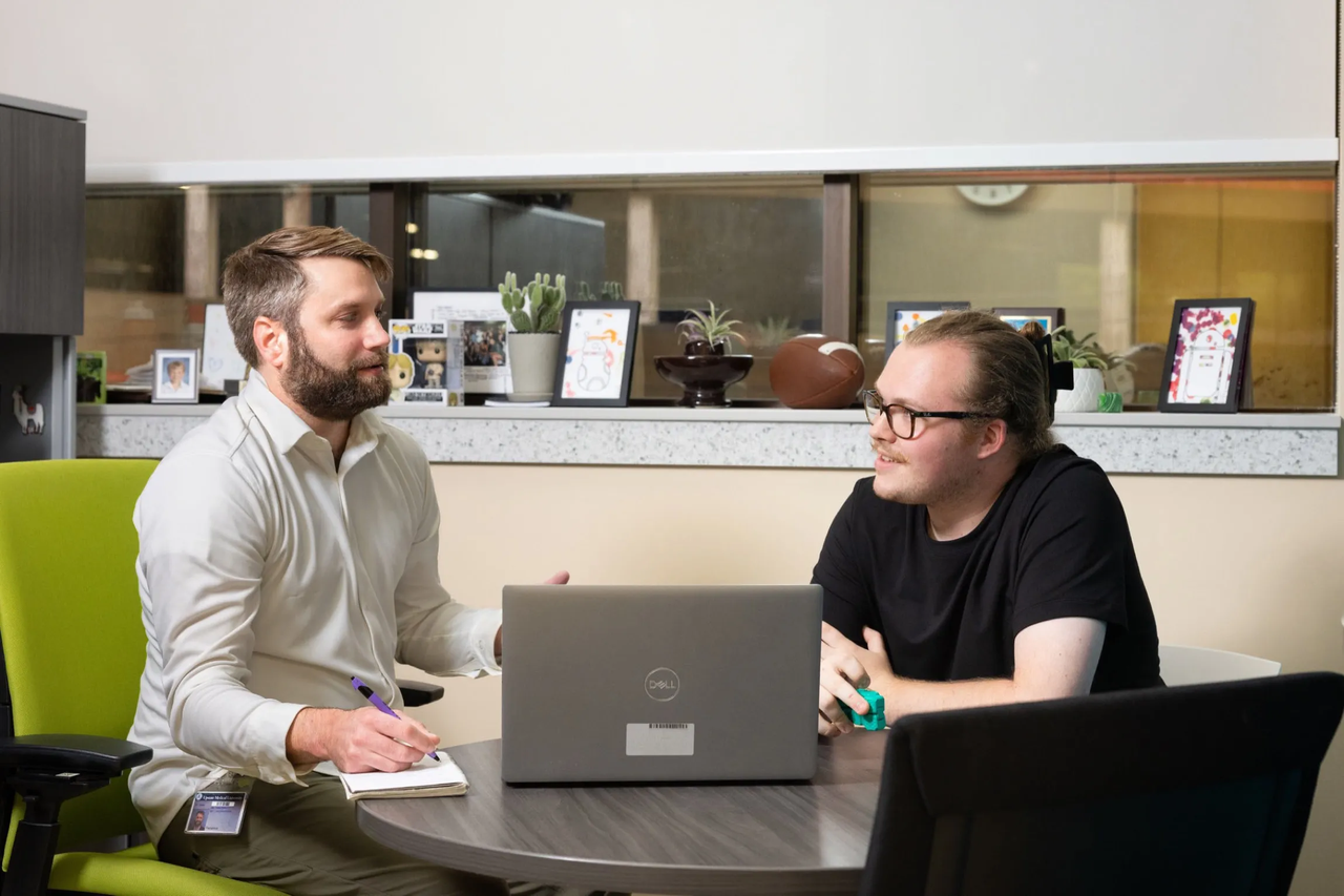 Two men speak seated at a table with a laptop open before them.