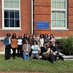 A group of 13 students take a group photo in front of Peck Hall.