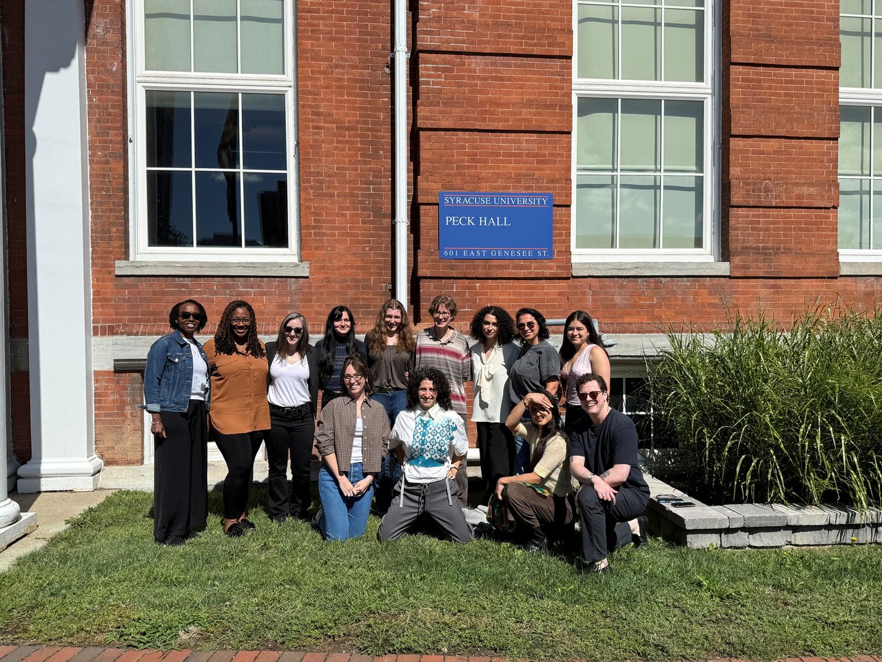 A group of 13 students take a group photo in front of Peck Hall.