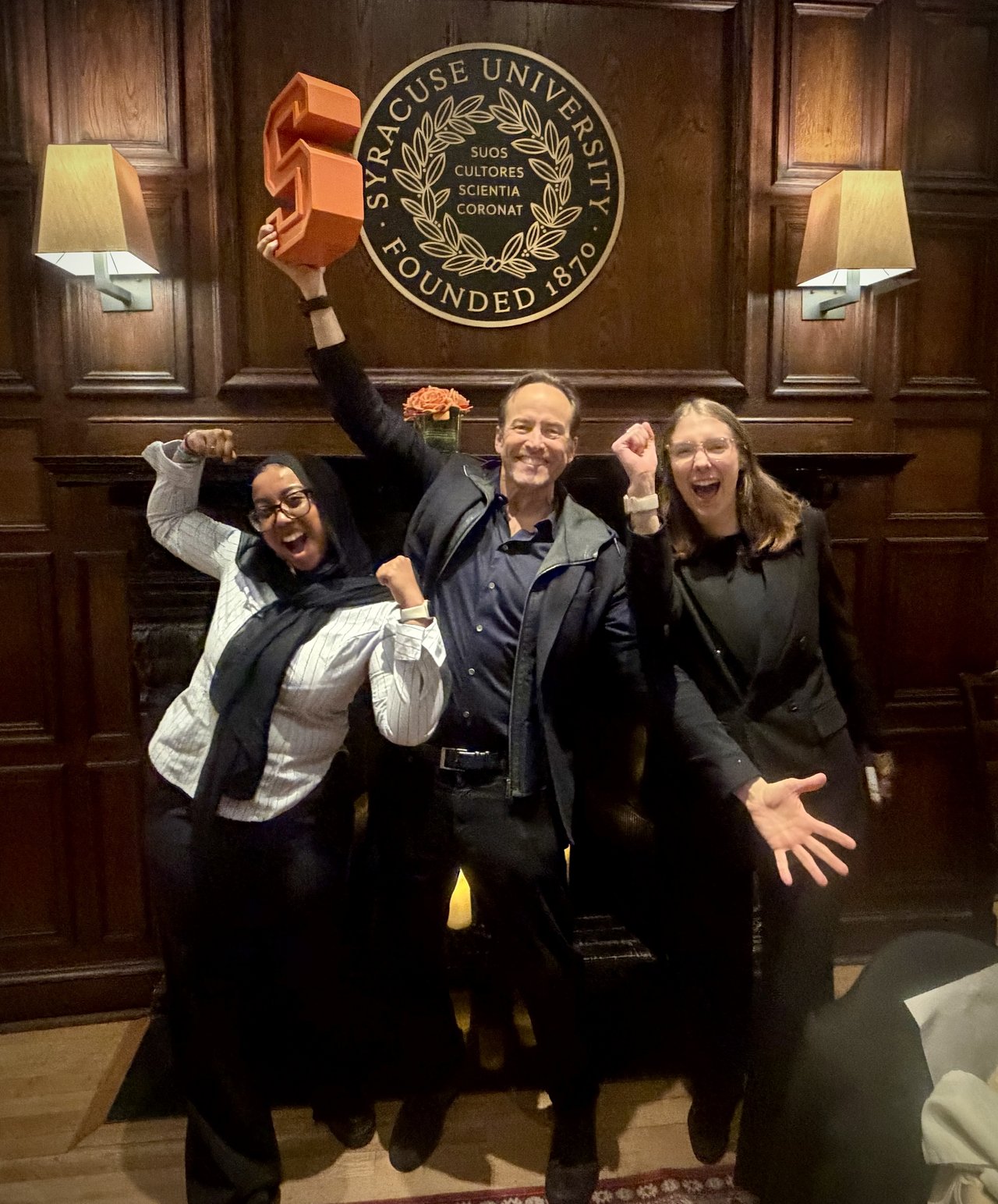 Three people celebrate exuberantly in front of a Syracuse University seal on a wood-paneled wall, with one person raising an orange block-letter 'S' trophy above their head.