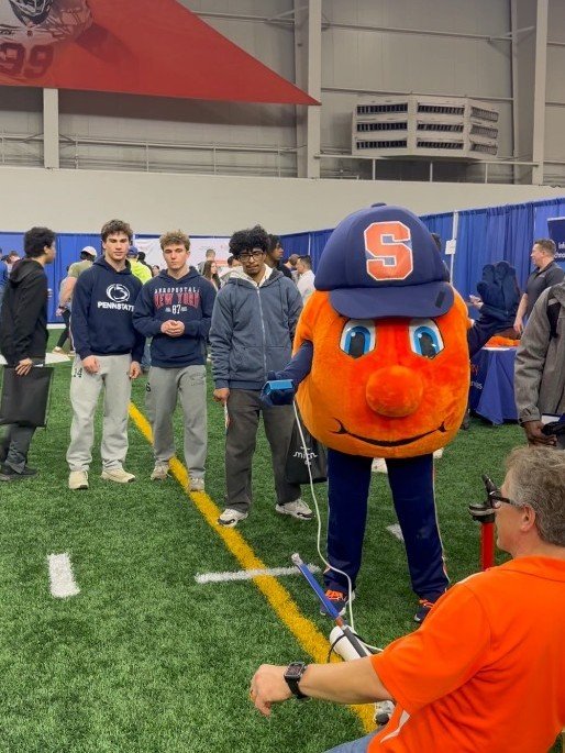 Otto the Orange, Syracuse University's mascot, stands on an indoor turf field surrounded by students at an event, while a person in an orange shirt operates equipment in the foreground.