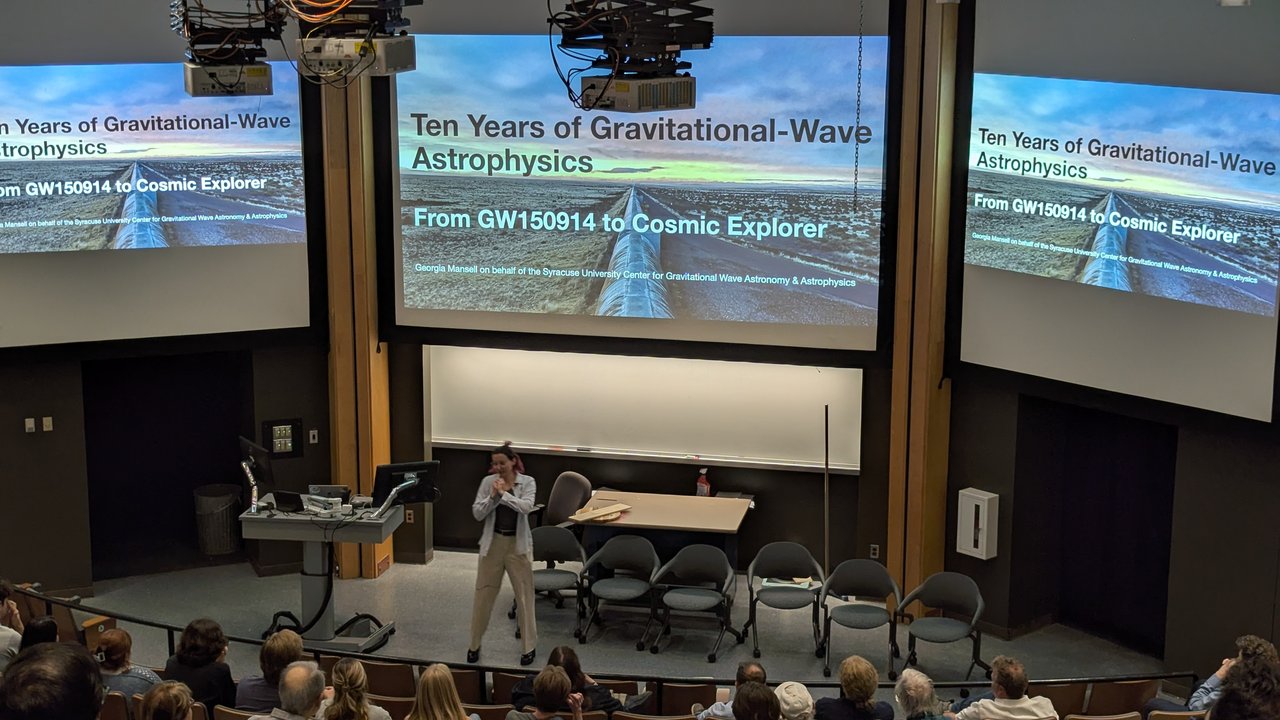 Person speaking to a room of people with three projector screens in the background.