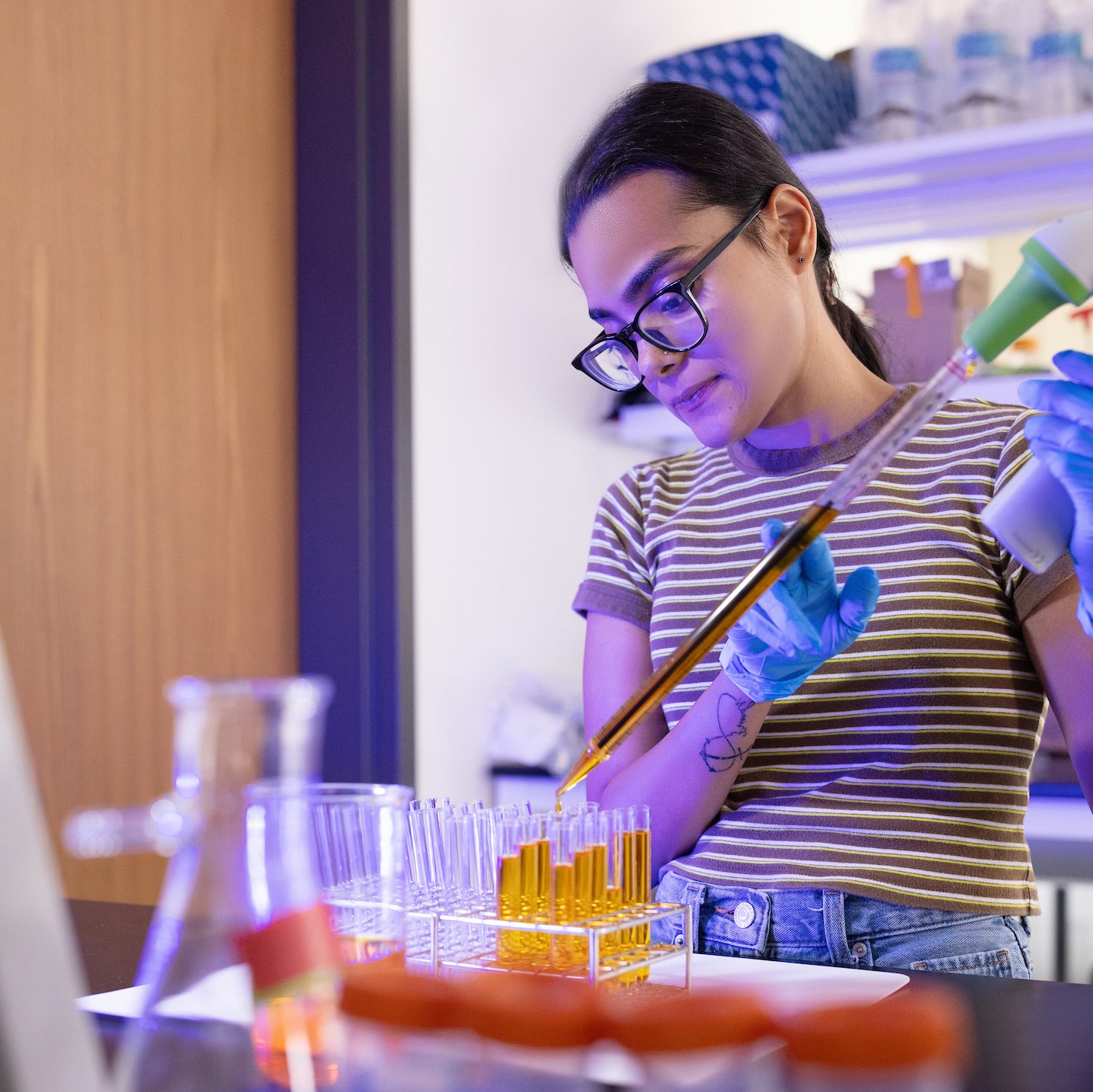 A young woman wearing glasses and blue latex gloves carefully pipettes a golden-colored liquid into a rack of test tubes in a modern, blue-lit laboratory setting.