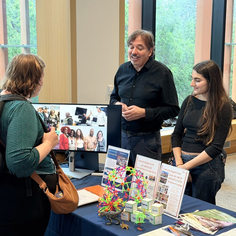 Students and staff talking at a university outreach table showcasing community‑engaged courses and student projects, with a monitor displaying group photos and colorful educational models, brochures, and toys arranged on the table.