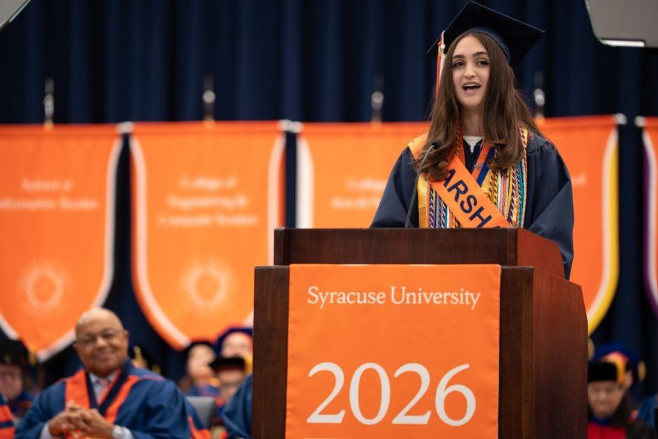 A graduate wearing a cap, gown, and orange Class Marshal sash speaks at a podium displaying 'Syracuse University 2026,' with orange school banners hanging behind her.