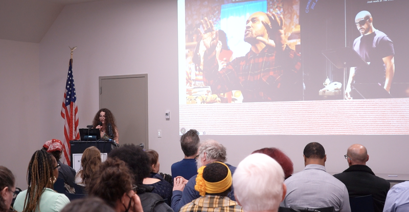 Person addressing a crowd in front of an image projected on a wall.