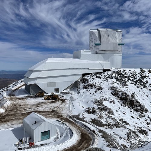 Rubin Observatory on a snowy mountaintop under a patchy blue sky with wispy clouds. The observatory is boot-shaped, with long white service building extending left and angular silver dome sticking up on the right. A small shed is visible to the left and in front of the observatory. A brown dirt road curves around the right side of the shed toward the observatory.