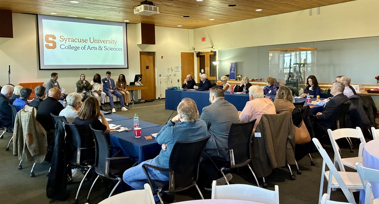 Group of people seated around tables in front of a panel speaking.