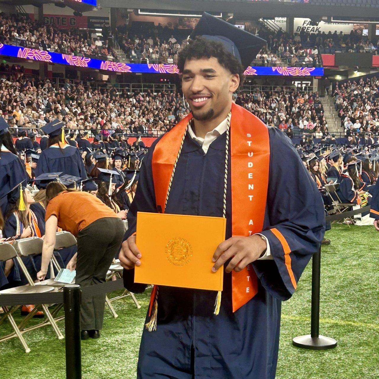 A smiling graduate in cap, gown, and an orange 'Student Athlete' stole holds up an orange Syracuse University diploma folder inside a packed stadium.