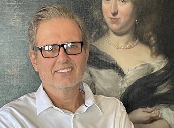 A man in glasses stands in front of a painting of a woman