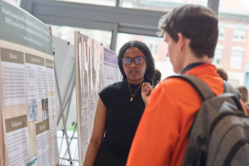 A student wearing glasses speaks to an attendee in an orange jacket at an academic poster session, with her research board visible behind her.