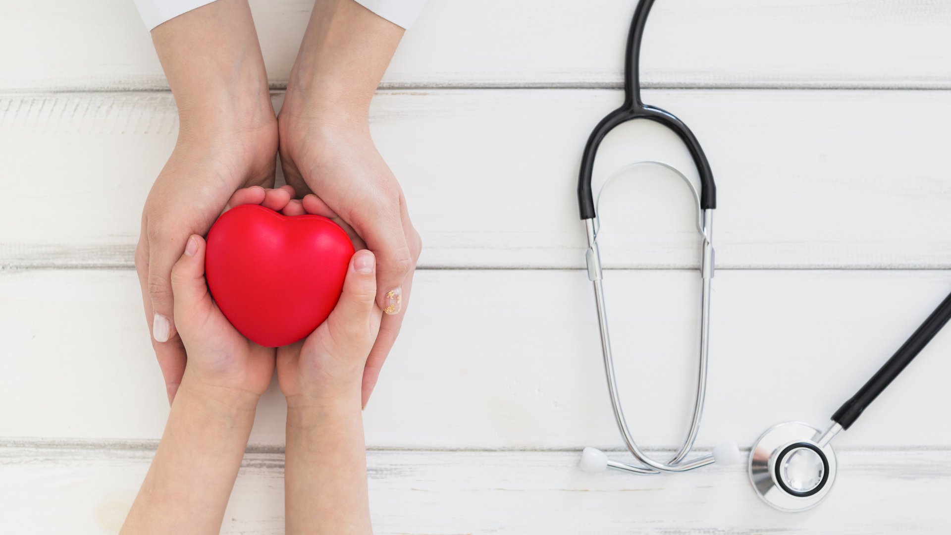 Two pairs hands holding a rubber heart next to a stethoscope.
