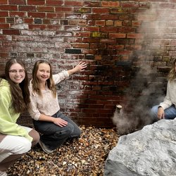 Three women bending down in front of a steam vent.
