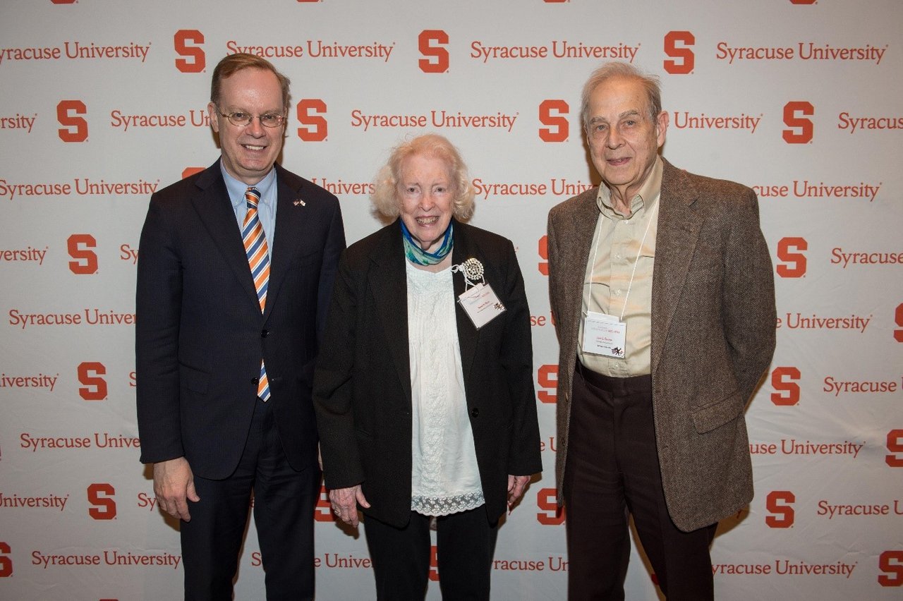 Three people pose for a photo in front of a Syracuse University backdrop.