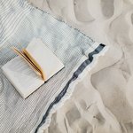 An open paperback book rests on a striped, fringed blanket spread out on sandy beach, with footprints visible in the sand nearby.
