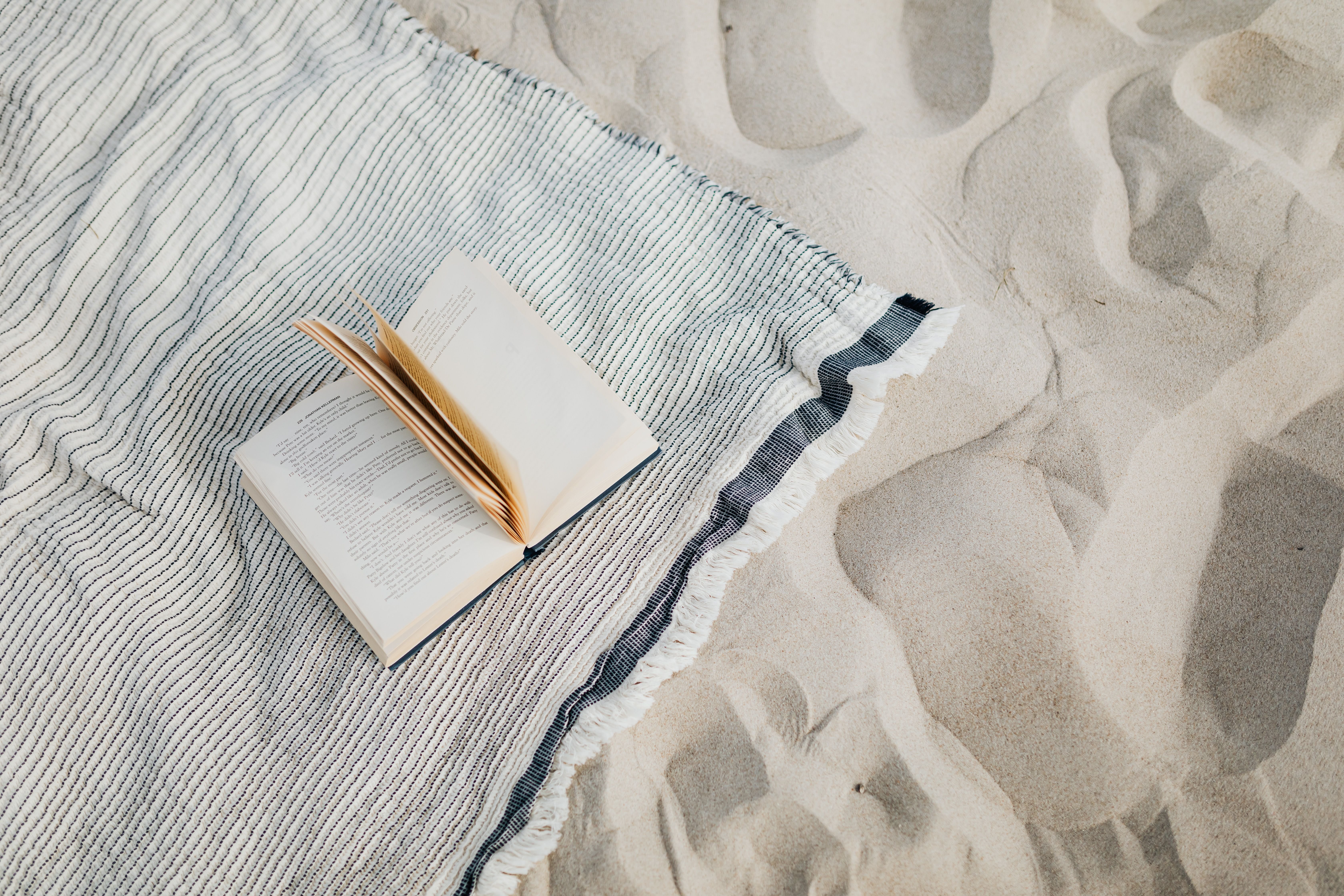 An open paperback book rests on a striped, fringed blanket spread out on sandy beach, with footprints visible in the sand nearby.